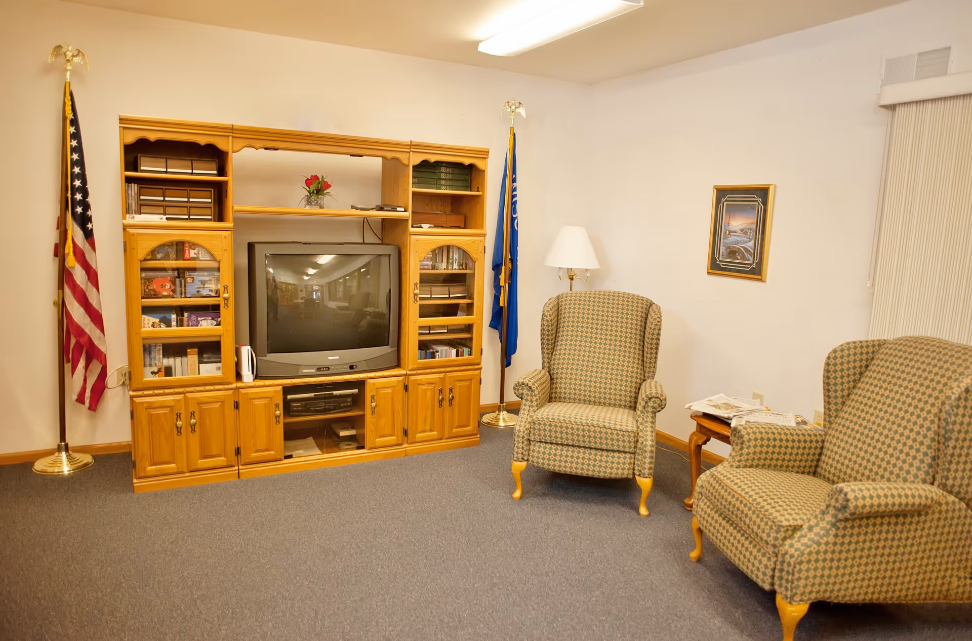 A cozy living room with two patterned armchairs, a wooden entertainment center holding a television, books, and decorative items. There are two flags on either side of the entertainment center, a floor lamp next to one armchair, a small wooden side table with newspapers, and a framed picture on the wall.