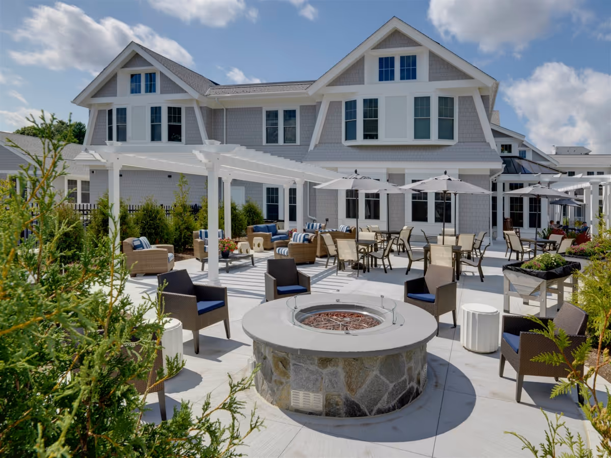 Outdoor patio area at The Residence at Penniman Hill featuring a stone fire pit surrounded by wicker chairs with blue cushions. There are multiple seating areas with tables, chairs, and umbrellas, as well as a white pergola and greenery around the patio. The building in the background has gray siding and white trim with multiple windows under a partly cloudy sky.