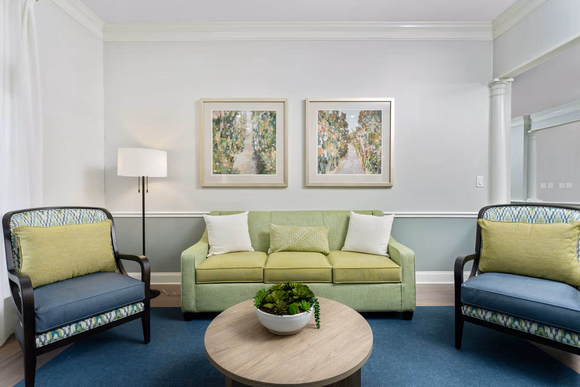 A cozy living room with a light green sofa adorned with three cushions, flanked by two armchairs with blue seats and green cushions. A round wooden coffee table with a small plant centerpiece sits in front of the sofa. Two framed paintings of floral scenes hang on the light gray wall above the sofa. A floor lamp stands to the left side of the sofa, and a large mirror with white columns is visible on the right side of the room.