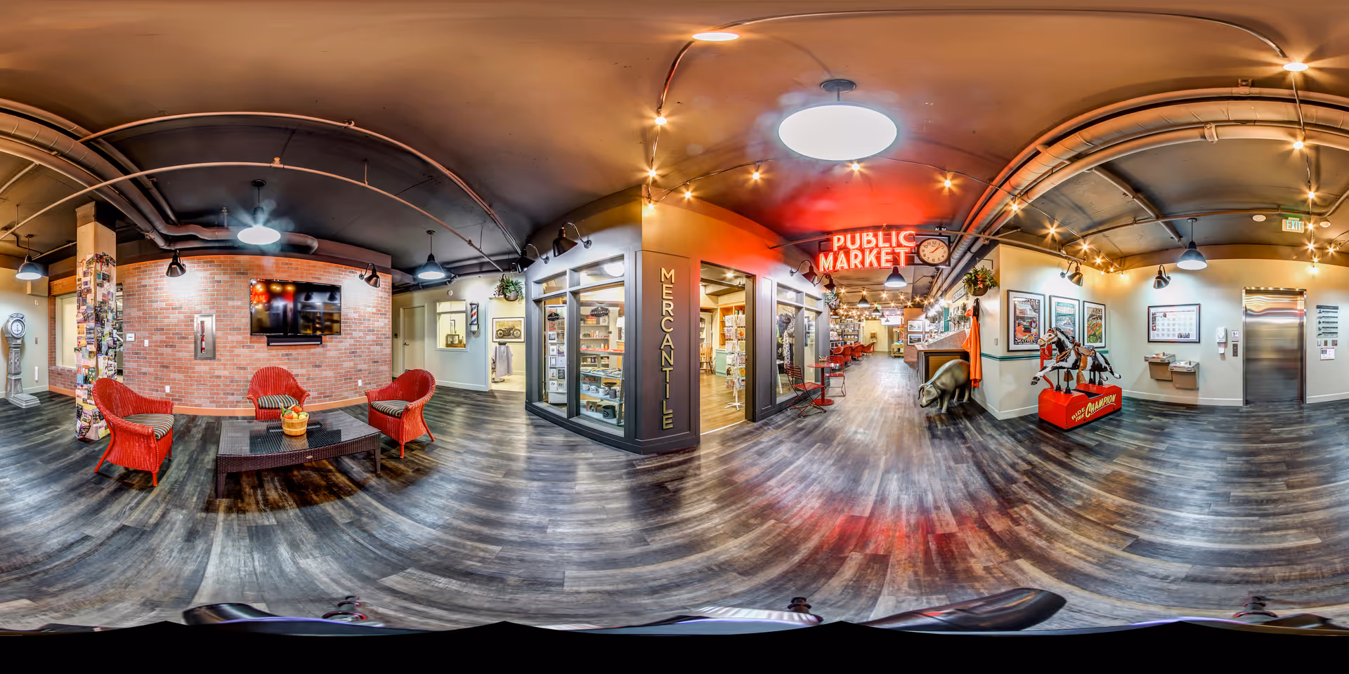 A spacious, well-lit interior common area with wood flooring and a mix of modern and vintage decor. The left side features a seating area with red wicker chairs and a coffee table in front of a brick wall with a mounted TV. The center has a storefront labeled 'Mercantile' with glass windows displaying various items. The right side shows a hallway with a neon sign reading 'Public Market' above, vintage-style decorations including a carousel horse, and framed pictures on the walls. The ceiling has exposed pipes and multiple hanging lights.
