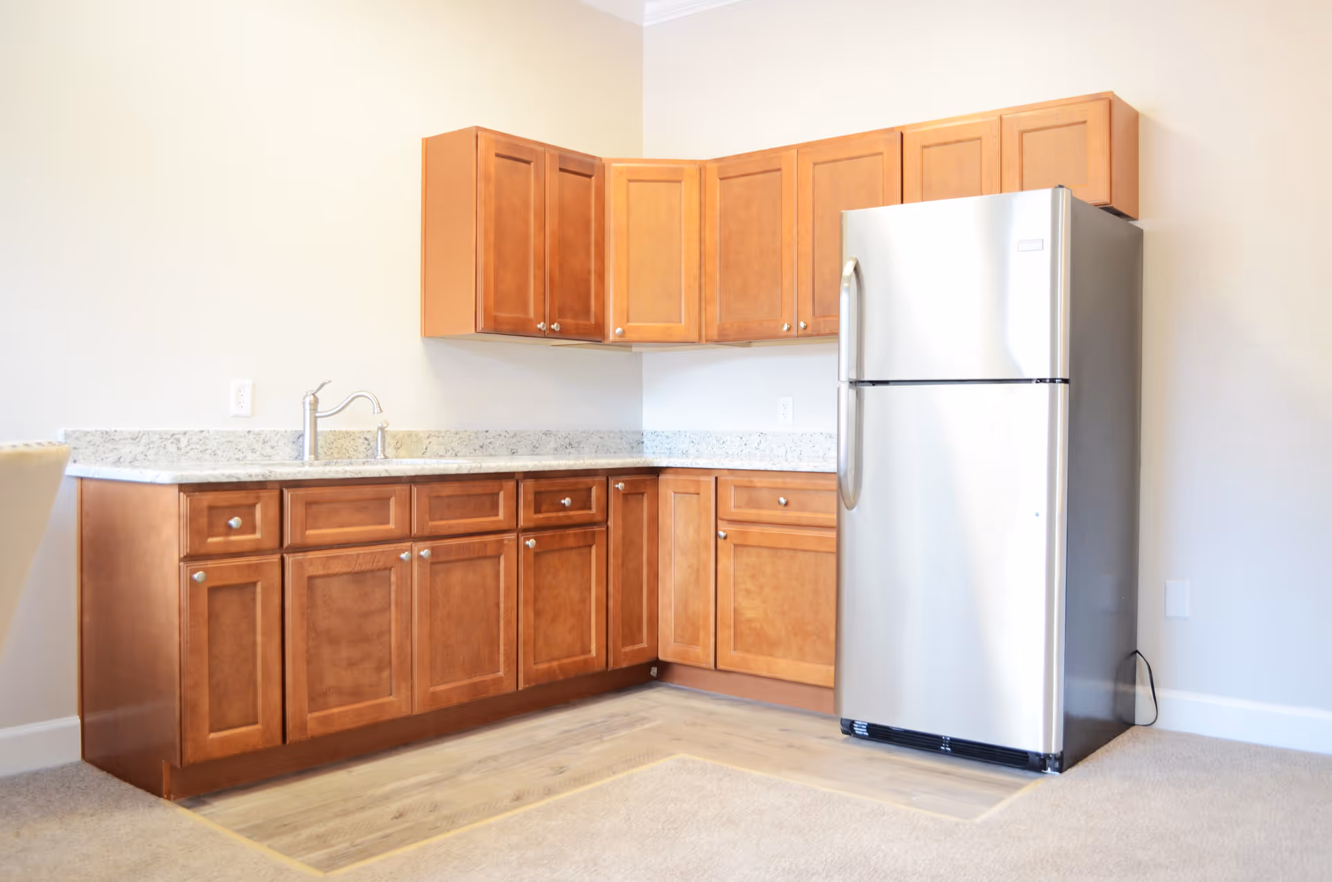 A kitchen area with wooden cabinets, a granite countertop, a stainless steel refrigerator, and a sink with a faucet. The floor transitions from carpet to wood near the kitchen space.