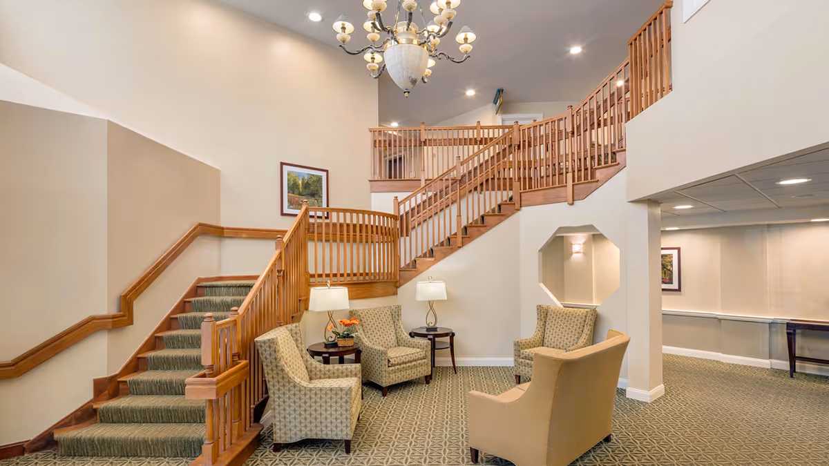 Interior lobby with a carpeted staircase and wooden railing, chandelier overhead, and a seating area with armchairs and lamps.