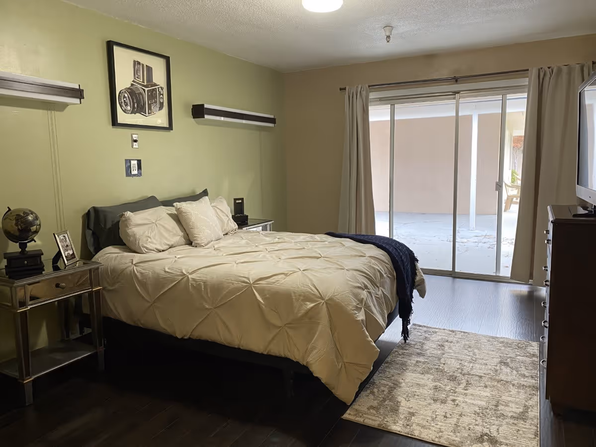 A bedroom with a neatly made bed featuring cream-colored bedding and multiple pillows. There is a small mirrored nightstand with a globe and picture frame on the left side of the bed. Above the bed is a framed picture of a vintage camera. The room has light green walls, dark wood flooring, and a sliding glass door with beige curtains that leads to an outdoor patio. A dresser with a TV on top is visible on the right side of the room.