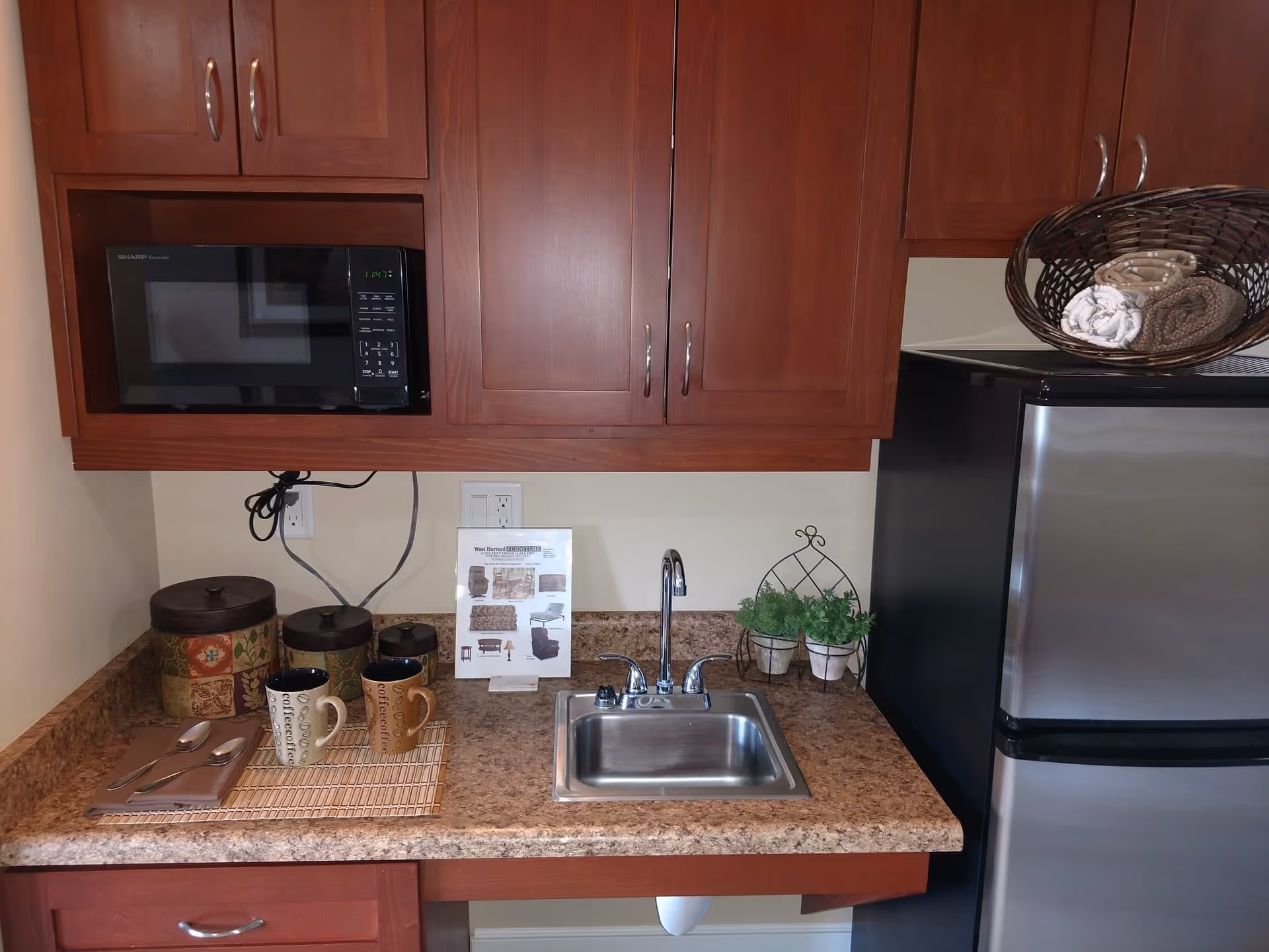 A small kitchen area with wooden cabinets, a black microwave, a stainless steel mini refrigerator, a small stainless steel sink, a countertop with two coffee mugs, three decorative canisters, a small plant in a wire holder, and a placard with furniture images.