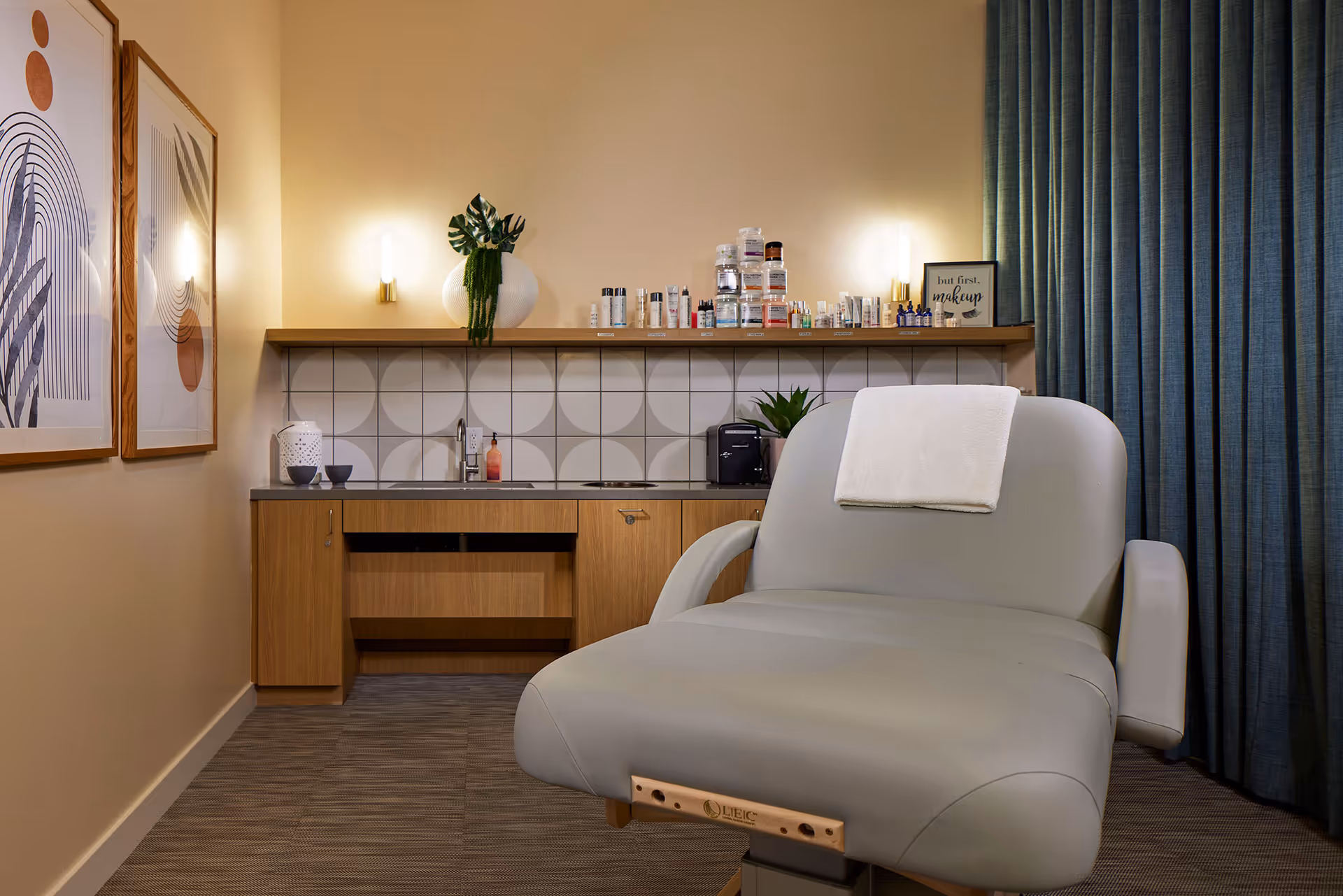 A modern and clean beauty or treatment room with a large gray reclining chair in the foreground, a wooden cabinet with a sink and various skincare products on a shelf above it, two framed abstract art pieces on the left wall, and blue curtains on the right side.