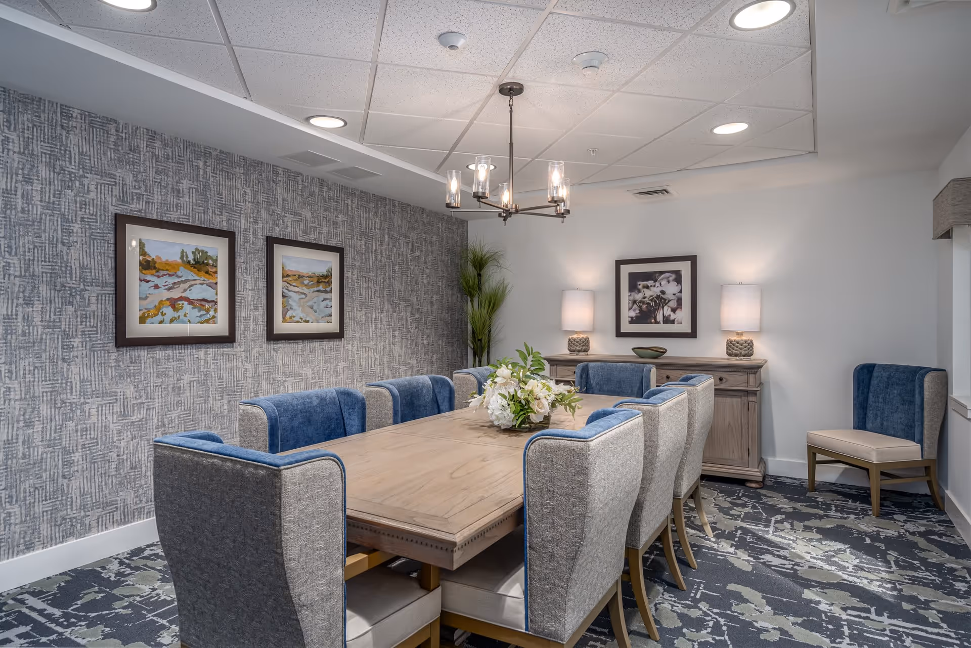 Well-lit dining room with a long wooden table surrounded by upholstered chairs, artwork on the walls, lamps, and a chandelier.