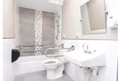 A clean, modern bathroom with a white toilet, white sink, and a bathtub with a shower. The bathtub has a vertical decorative tile strip and multiple grab bars for accessibility. There is a mirror above the sink and a soap dispenser mounted on the wall.