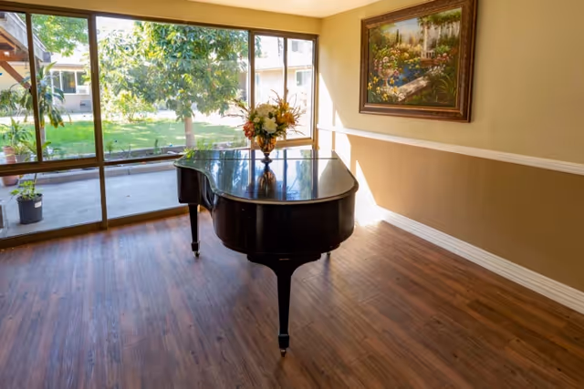 A bright room with wooden flooring featuring a black grand piano with a floral arrangement on top. Large windows provide a view of a green outdoor area with trees and plants. A framed painting hangs on the beige wall.