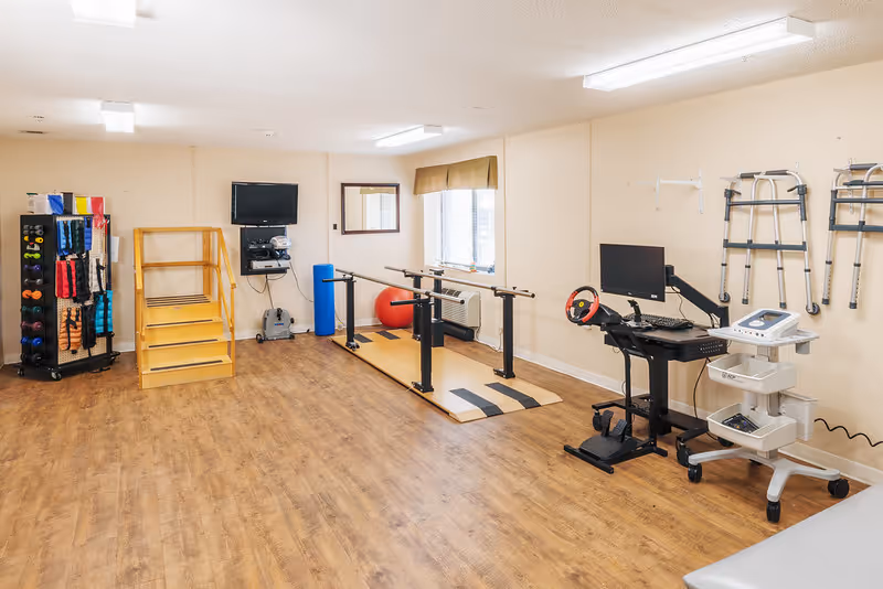 Rehabilitation room with parallel bars on a wood floor, exercise equipment and weights, walkers on the wall, and a TV/computer station.