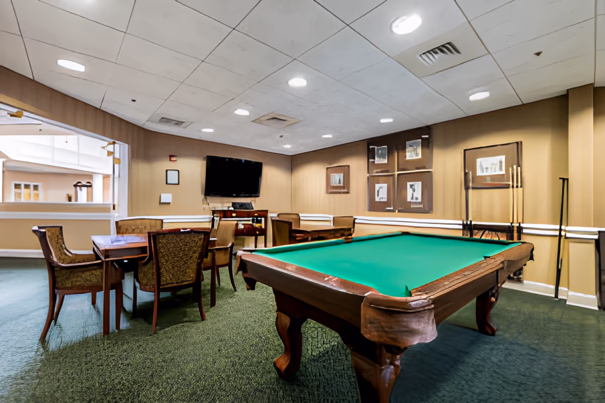 Interior view of a recreational room with a green felt pool table in the foreground, a set of chairs and tables to the left, a wall-mounted flat screen TV, and framed pictures on the walls. The room has green carpet and beige walls with white trim.