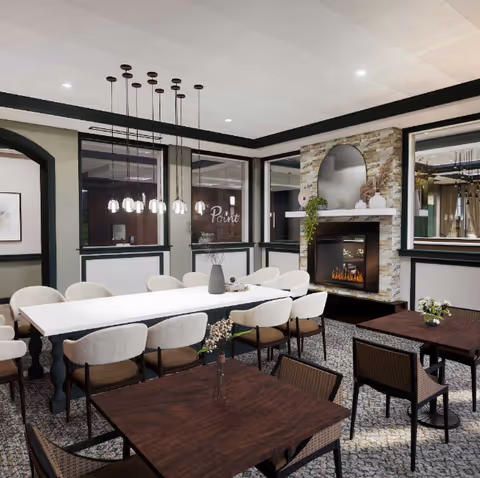 A cozy dining area in a senior living facility featuring a long white table surrounded by cushioned chairs, smaller dark wood tables with woven chairs, a stone fireplace with a mirror above it, and pendant lights hanging from the ceiling. Large windows and neutral-colored walls create a warm and inviting atmosphere.