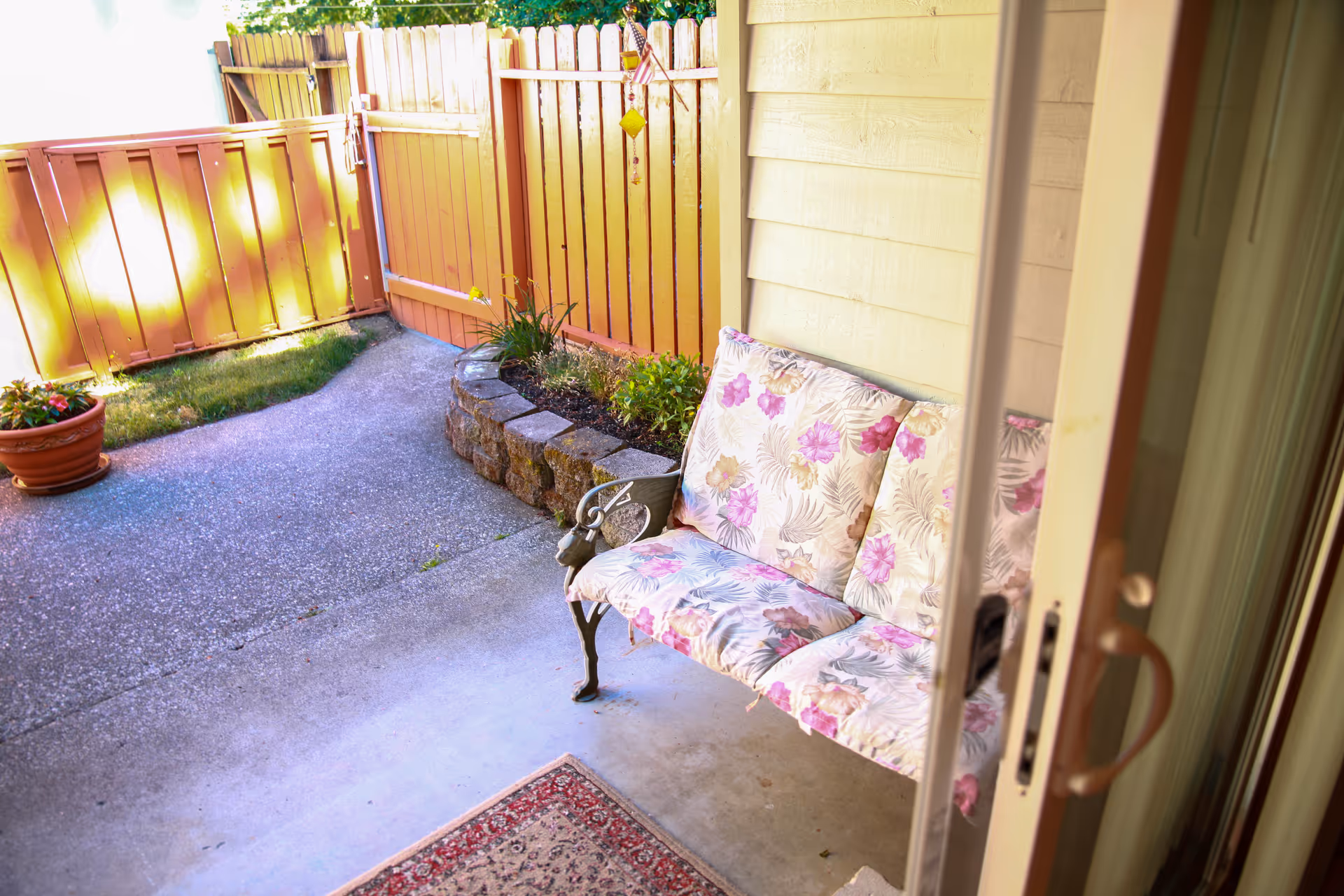 A small outdoor patio area with a floral cushioned bench, a potted plant, a curved stone planter with greenery, and a wooden fence with a gate. The patio is viewed through a partially open sliding glass door.