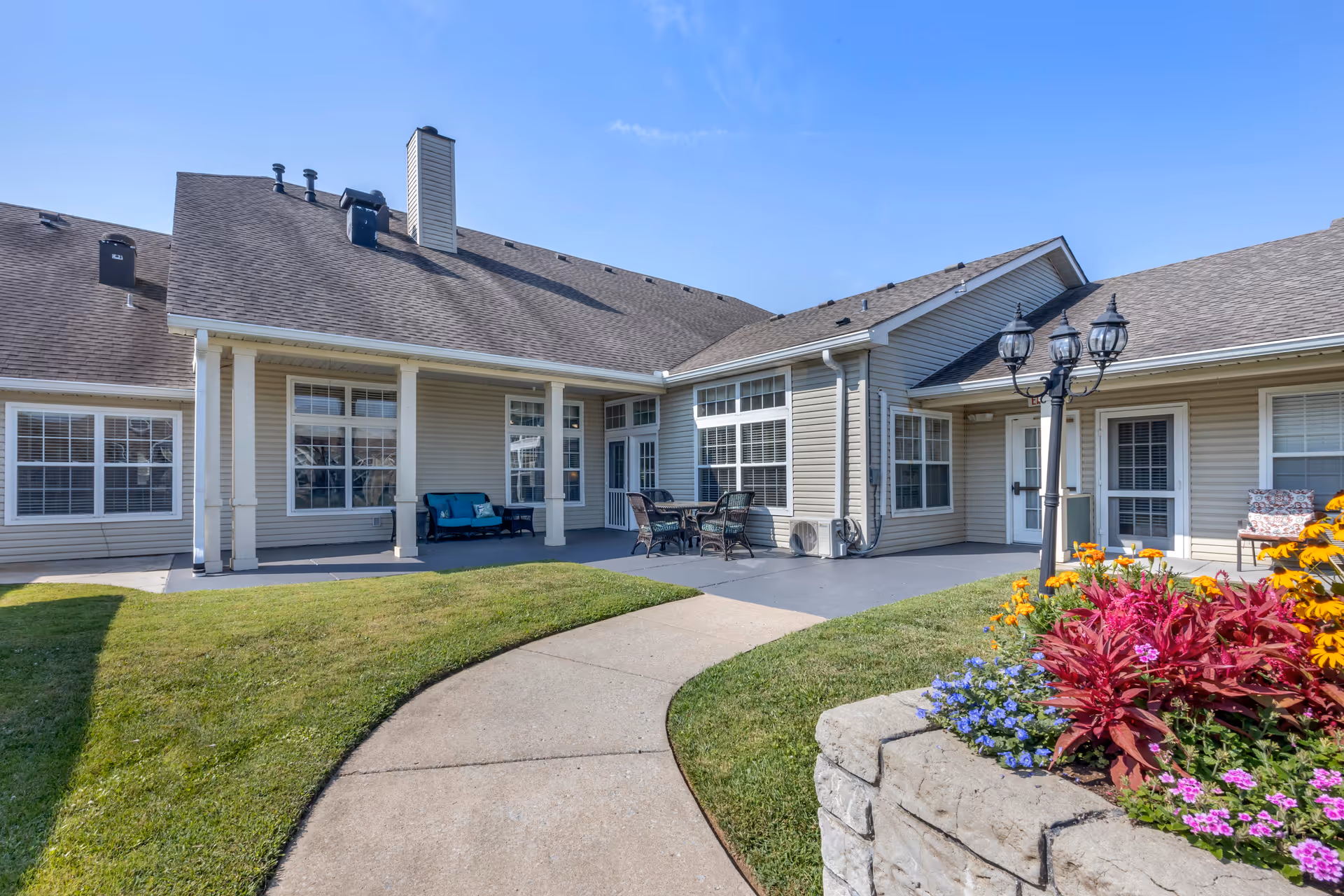 Outdoor patio area at Brookdale Gallatin featuring a curved concrete walkway, green lawn, a flower bed with colorful flowers, a lamp post with three lanterns, and patio furniture including a cushioned bench and a table with four chairs. The building exterior has beige siding and multiple windows under a clear blue sky.