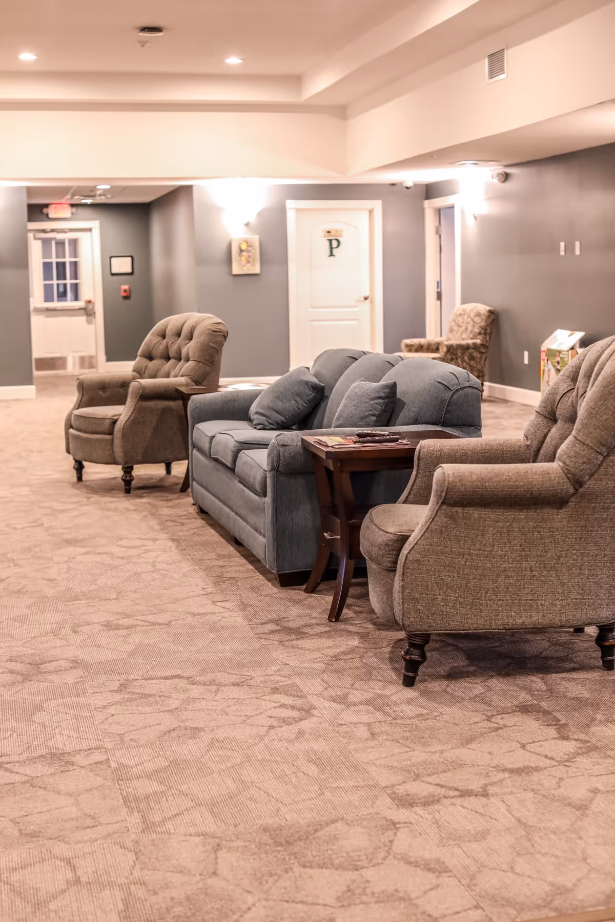 A cozy seating area in an assisted living facility hallway with a blue cushioned sofa, two beige armchairs, a wooden side table with magazines, and soft lighting on the walls. The carpet has a patterned design and there are doors and a window visible in the background.