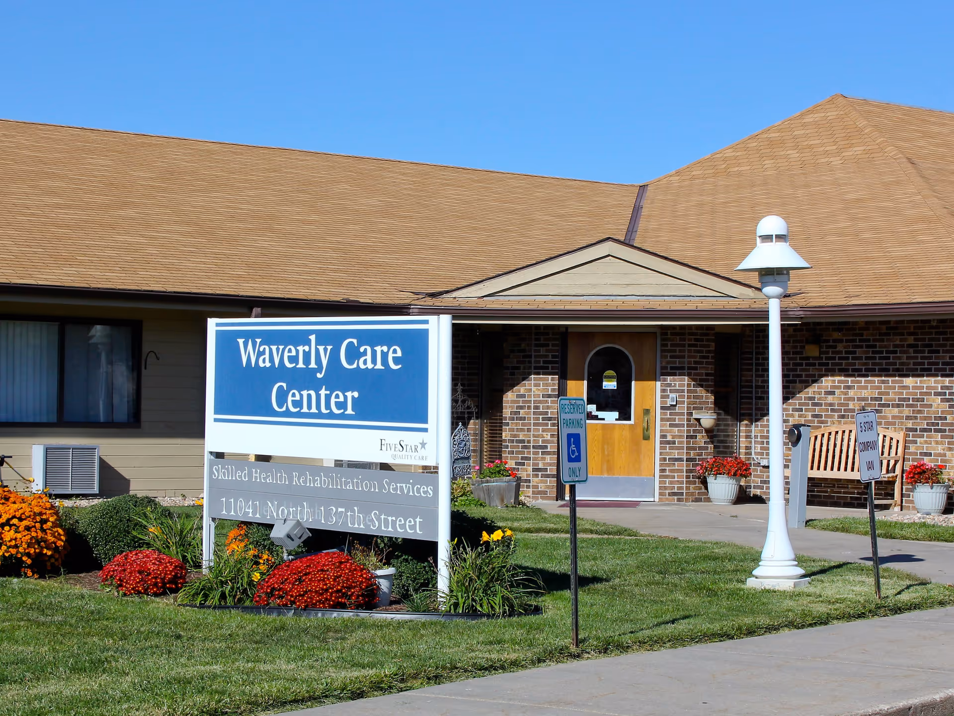 Front exterior of the Waverly Care Center showing the entrance, a large sign, flowers, and a lamp post.