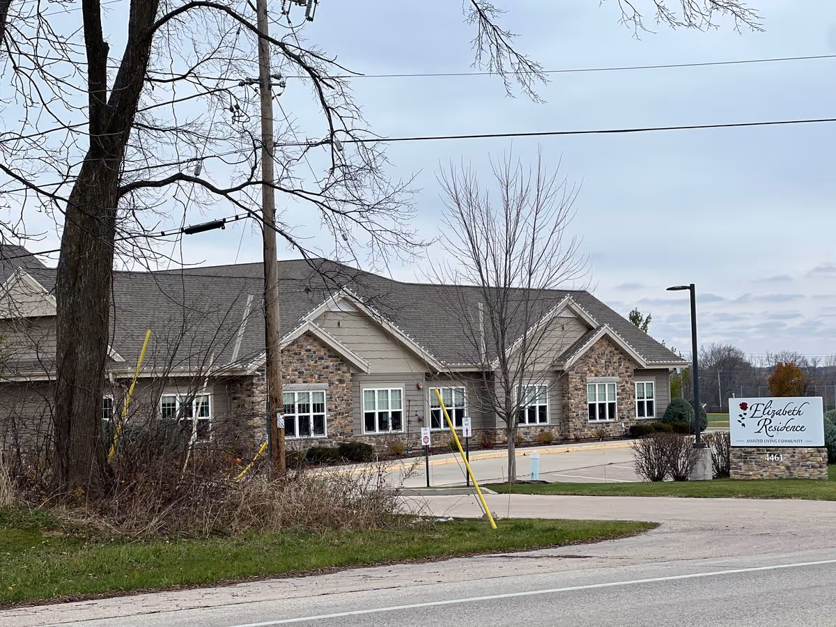 Front exterior of a single-story stone-and-siding senior living building with landscaping and an 'Elizabeth Residence' sign by the driveway.