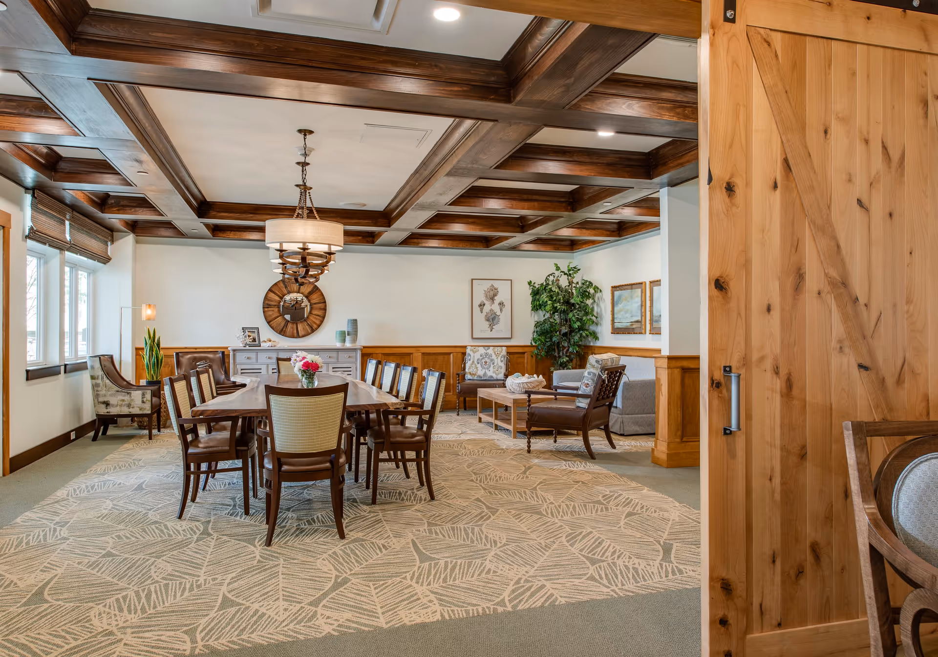 A spacious, well-lit dining and sitting area with a long wooden dining table surrounded by chairs. The room features a coffered wooden ceiling, a large chandelier, patterned carpet, and a mix of armchairs and a sofa near the back wall. There are windows on the left side letting in natural light, and decorative elements including framed pictures, a round mirror, plants, and a wooden sliding door partially open on the right.