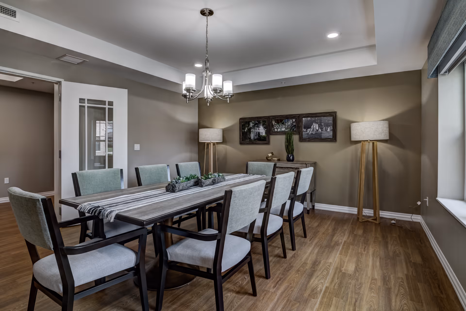 A dining room with a long wooden table surrounded by upholstered chairs, an overhead chandelier, two floor lamps, and wall art.