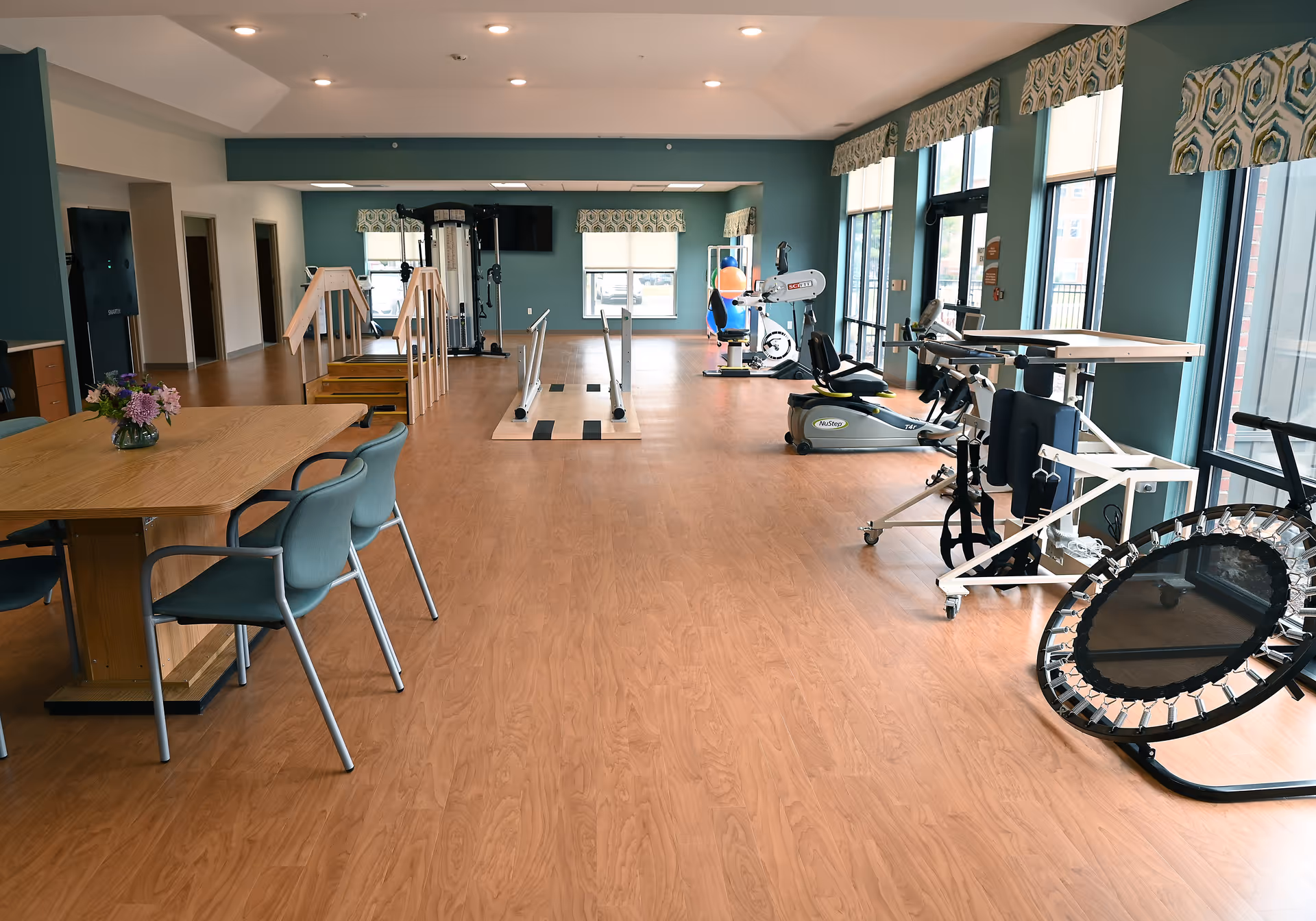 A spacious rehabilitation room with wooden flooring, featuring exercise equipment including a stationary bike, parallel bars, a small staircase for physical therapy, and a trampoline. There is a wooden table with chairs on the left side and large windows with patterned valances letting in natural light.