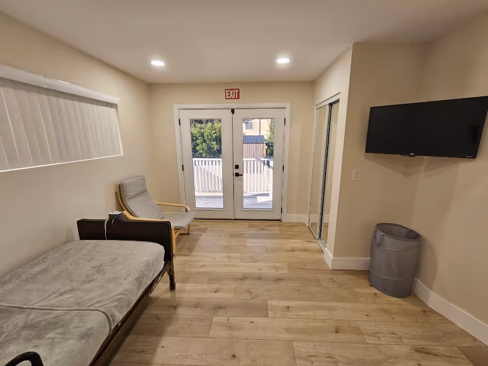 A simple room with light wood flooring, a single bed with a gray blanket on the left, a gray cushioned chair next to the bed, a wall-mounted flat-screen TV on the right, a gray laundry basket below the TV, and double glass doors with an exit sign above them leading outside.