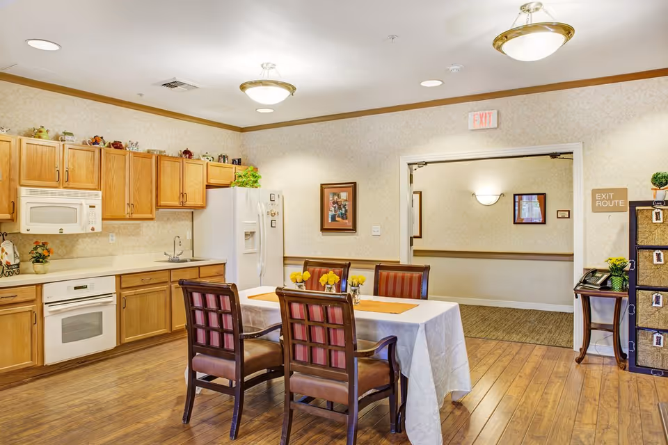 A bright kitchen and dining area in a senior living facility with wooden cabinets, a white refrigerator, microwave, and oven. A rectangular table covered with a white tablecloth is surrounded by four wooden chairs with red and brown cushions. Yellow flowers in small vases decorate the table. The room has wooden flooring and beige patterned wallpaper. An exit route sign and a small table with a phone and decorative items are visible near the doorway leading to a hallway.