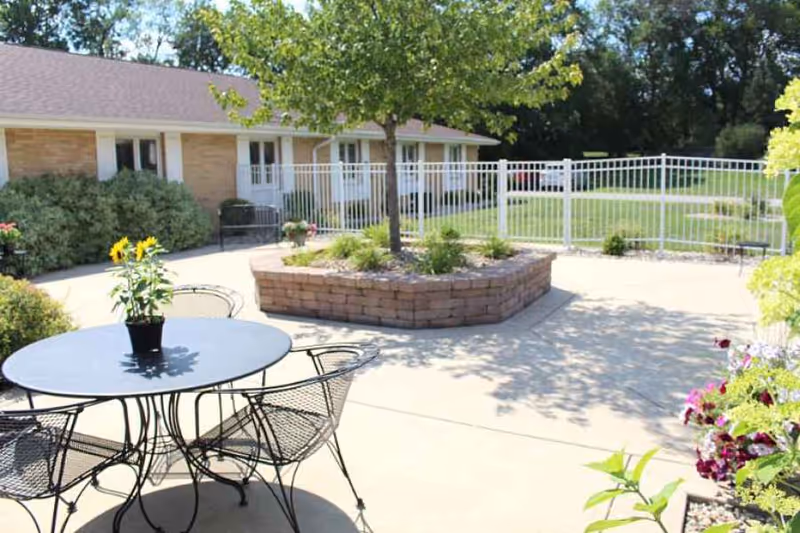 Outdoor patio area with a round metal table and four matching chairs. A small potted plant with yellow flowers is on the table. In the background, there is a raised brick planter with a tree and various plants. A white metal fence encloses the area, and a single-story building with beige brick walls and white doors and windows is visible behind the fence. The scene is bright and sunny with shadows cast on the concrete ground.