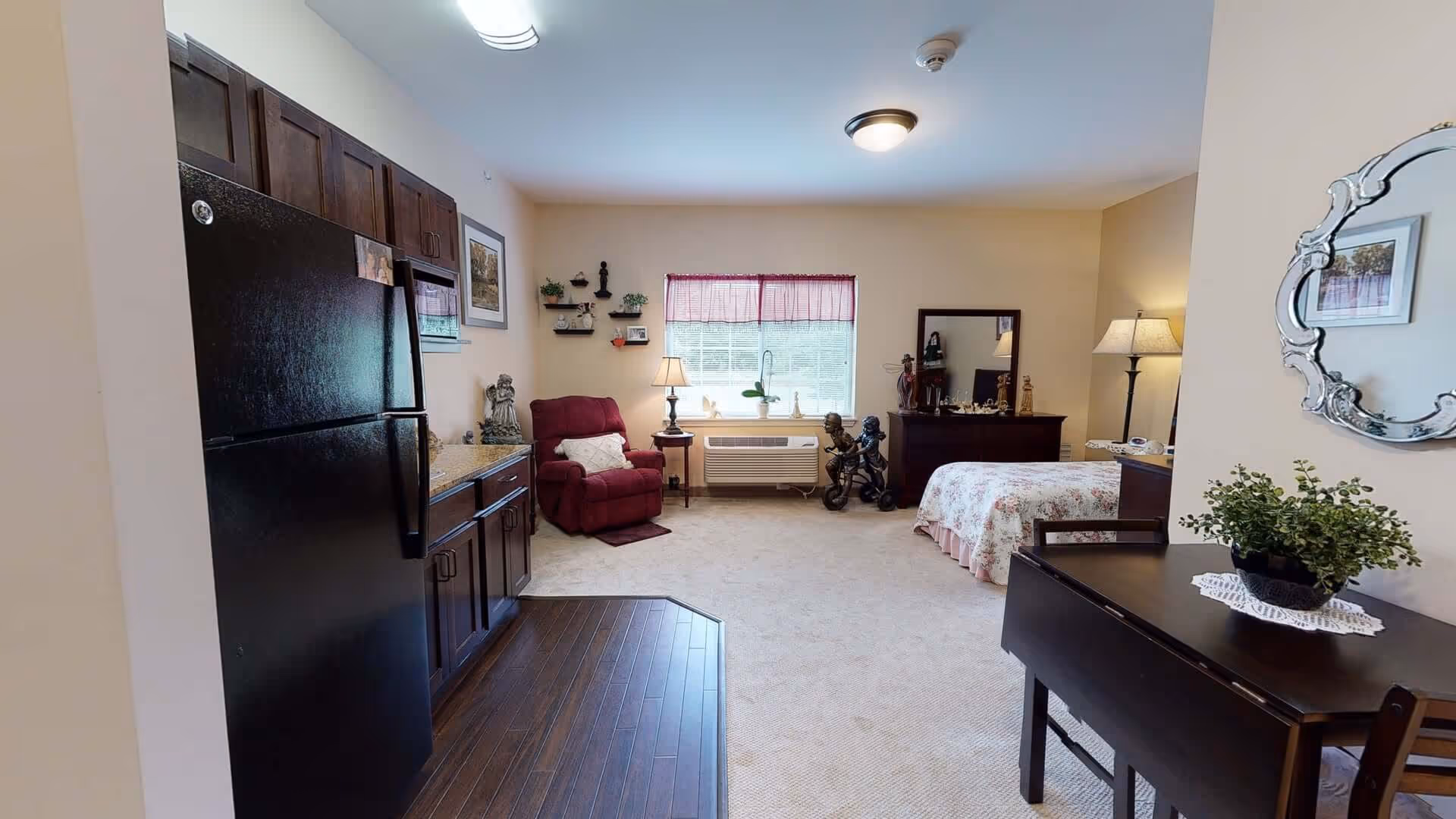 Sunlit studio apartment interior with a kitchenette and black refrigerator on the left, a recliner and bed in the center background, and a small table with a plant on the right.