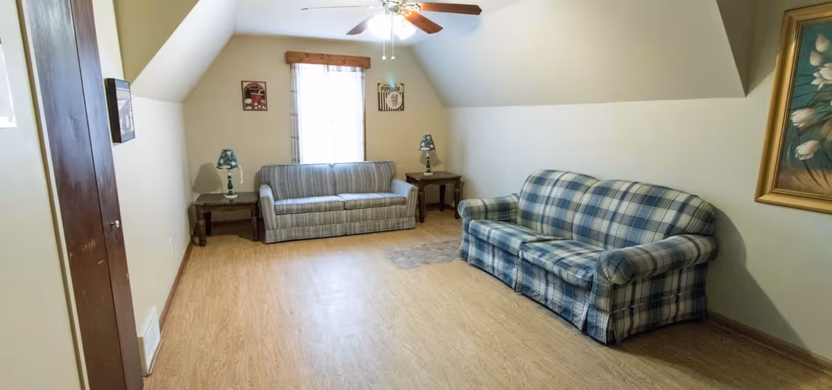 A small living room with two patterned sofas, one striped and one plaid, positioned against opposite walls. There are two wooden side tables with matching lamps beside each sofa. A window with a wooden frame and light curtains is centered on the far wall. The room has light-colored walls, a slanted ceiling, wood flooring, and a framed floral painting on the right wall.