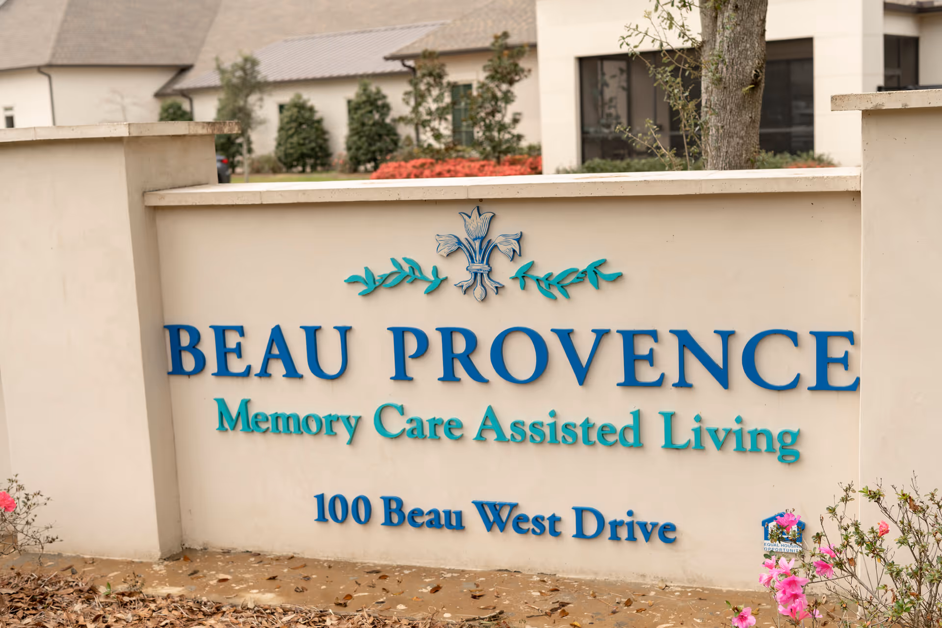Outdoor sign for Beau Provence Memory Care Assisted Living located at 100 Beau West Drive, with a decorative floral emblem above the text and some pink flowers and greenery around the base of the sign.