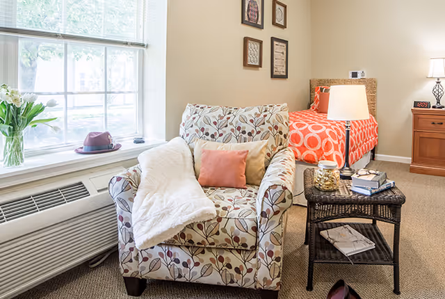 A cozy assisted living room featuring a floral patterned armchair with a white throw blanket and a pink cushion. Next to the chair is a wicker side table holding a lamp, books, and a jar of snacks. In the background, there is a bed with an orange and white circular patterned bedspread, a wooden nightstand with a lamp, and framed artwork on the wall. A large window with blinds lets in natural light.