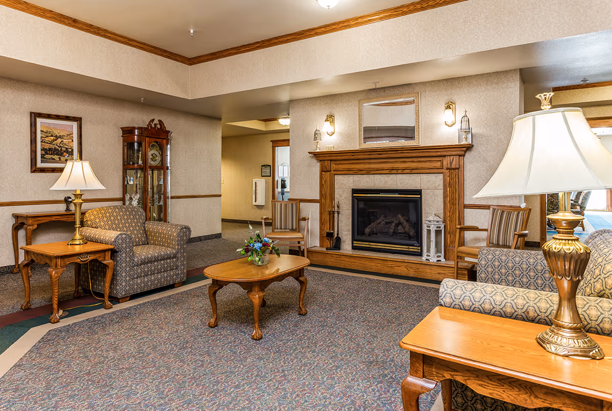 A cozy living room area in a senior living facility featuring patterned armchairs, wooden side tables with lamps, a wooden coffee table with a flower arrangement, and a fireplace with a wooden mantle. The room has beige walls with wooden trim, a carpeted floor, a framed landscape painting, and a glass-fronted curio cabinet.