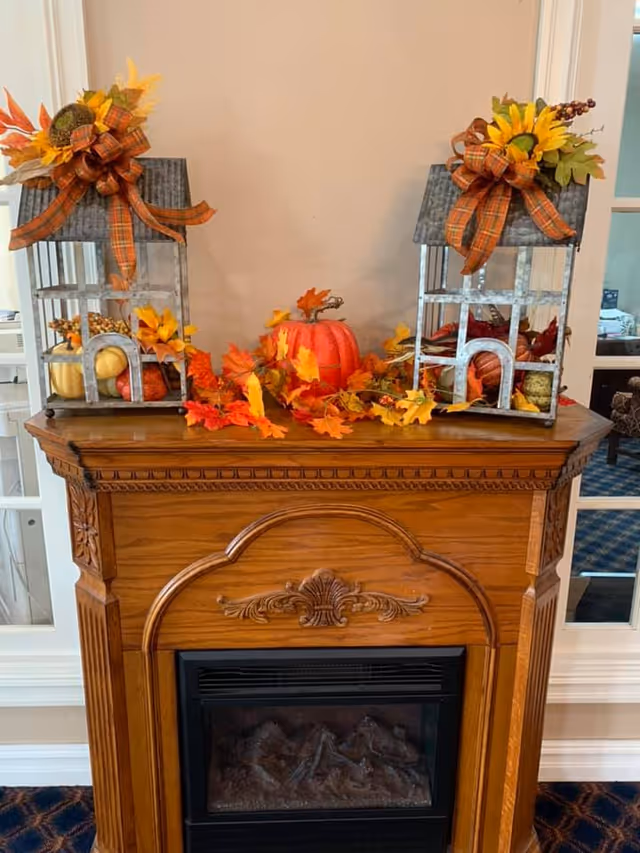 A wooden electric fireplace mantel decorated with autumn-themed items including two metal lanterns adorned with plaid bows and artificial sunflowers, a small orange pumpkin, and scattered fall leaves in shades of orange and yellow. The fireplace is set against a beige wall with white trim and a window with glass panes is visible on the right side.