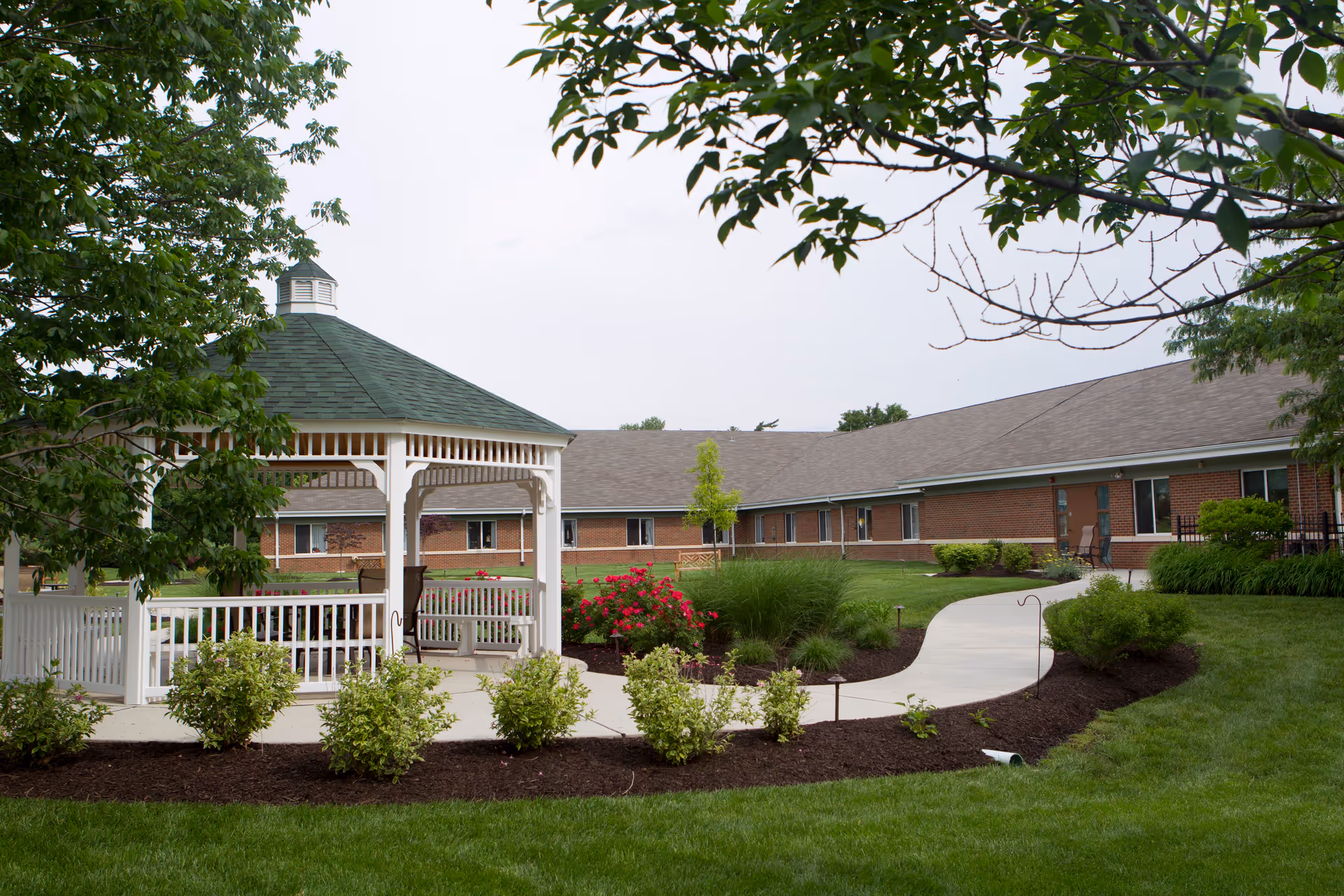 A landscaped outdoor area at Nazareth Living Center featuring a white gazebo with a green roof, surrounded by bushes, flowers, and a curved concrete walkway. The background shows a single-story brick building with multiple windows and a gray shingled roof.
