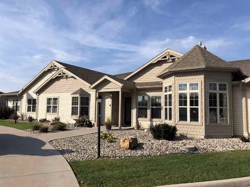 Exterior view of a single-story beige building with multiple windows and a small covered entrance. The building is surrounded by a landscaped area with rocks, shrubs, and a green lawn under a partly cloudy blue sky.