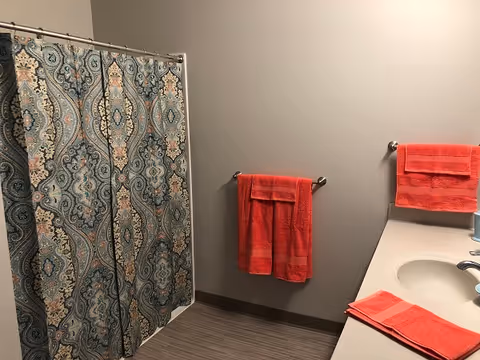 Bathroom with a patterned shower curtain, gray walls, countertop sink, and bright red towels hanging and folded on the counter.