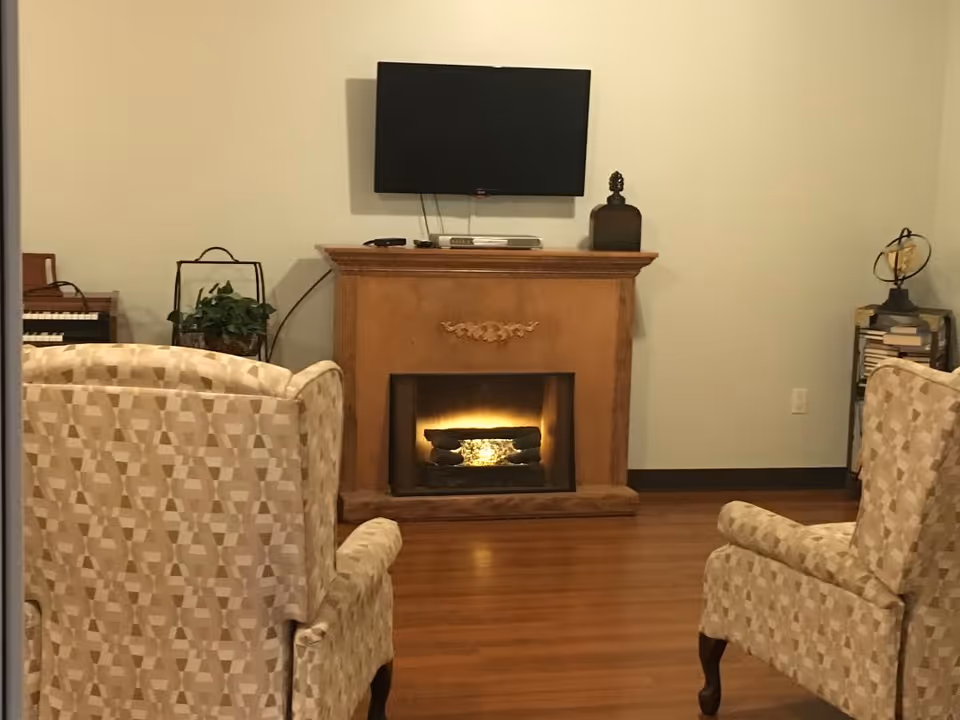 A cozy living room with two patterned armchairs facing a wooden fireplace with a glowing fire. Above the fireplace is a mounted flat-screen TV. To the left, there is a small organ and a plant, and to the right, a small bookshelf with books and a decorative globe on top. The room has wooden flooring and light-colored walls.