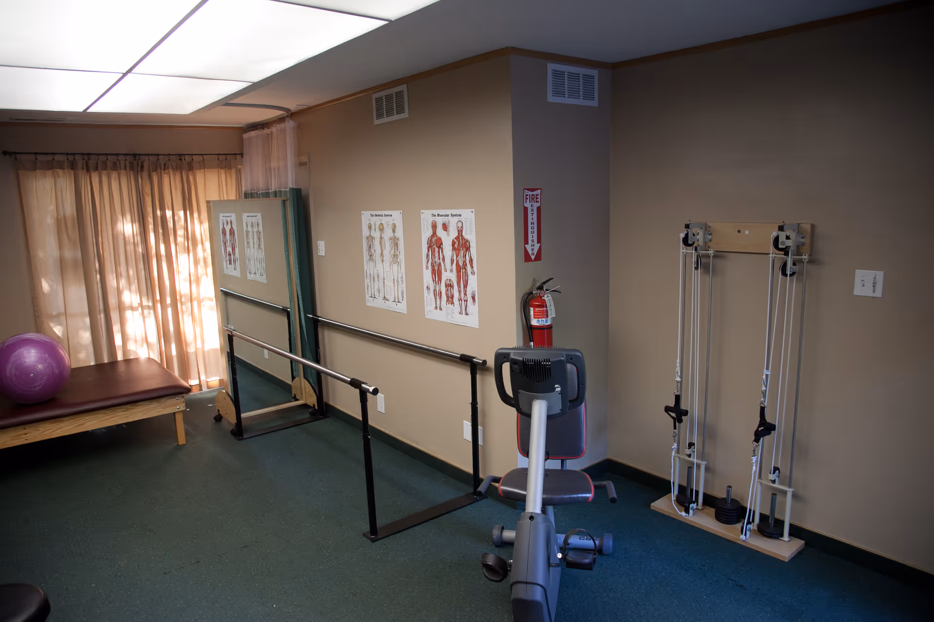 A small exercise room with a recumbent exercise bike, parallel bars for walking support, anatomical charts on the wall, a purple exercise ball on a padded bench, and a pulley weight system against the wall. The room has beige walls, green carpet, and a window covered with sheer curtains.