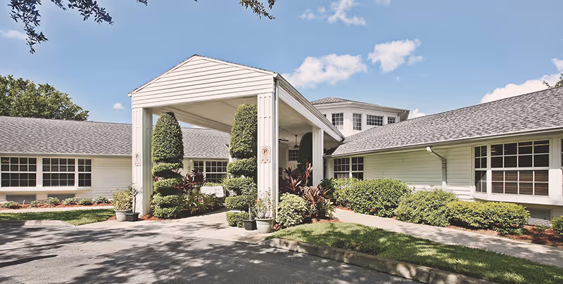 Front entrance of a single-story white senior living building with a covered porte-cochere and landscaped grounds.