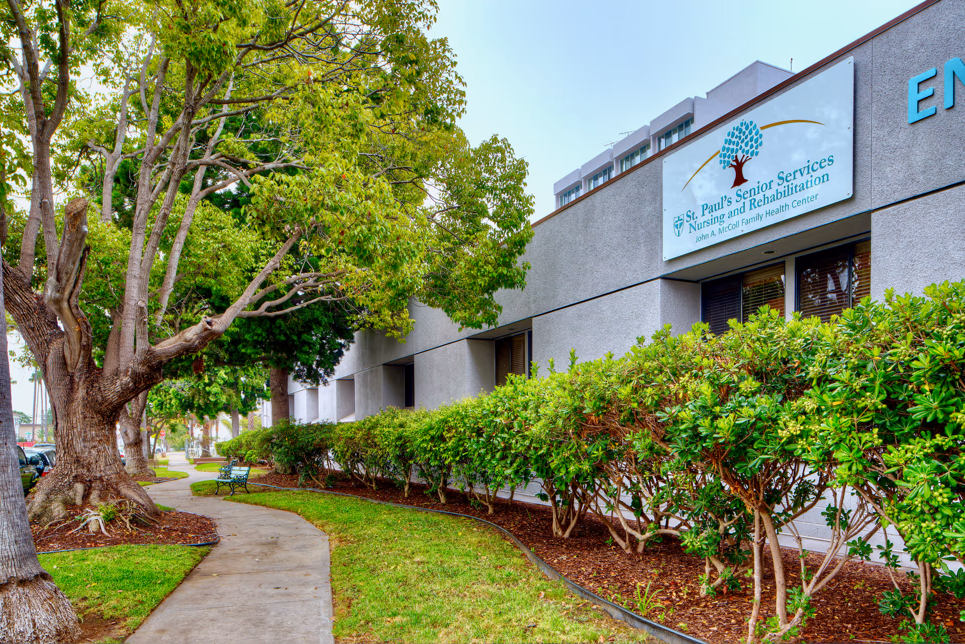 Landscaped sidewalk and exterior front of St. Paul's Skilled Nursing and Rehabilitation building with trees, shrubs, benches, and a visible facility sign.