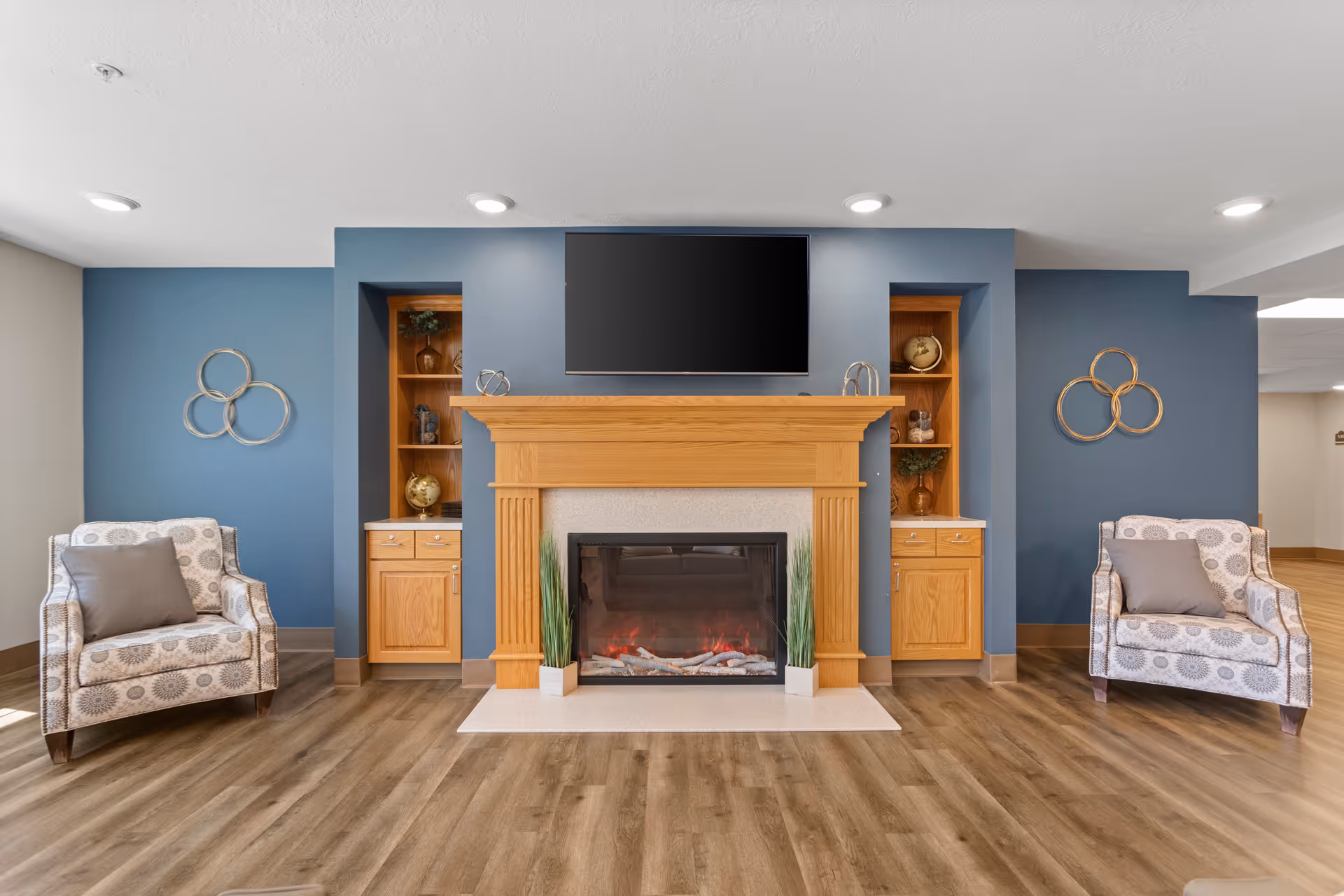 A cozy living room area with a wooden fireplace in the center, flanked by built-in wooden shelves and cabinets. Above the fireplace is a flat-screen TV mounted on a blue accent wall. On either side of the fireplace are two patterned armchairs with gray pillows. The floor is wood, and the room is well-lit with recessed ceiling lights.