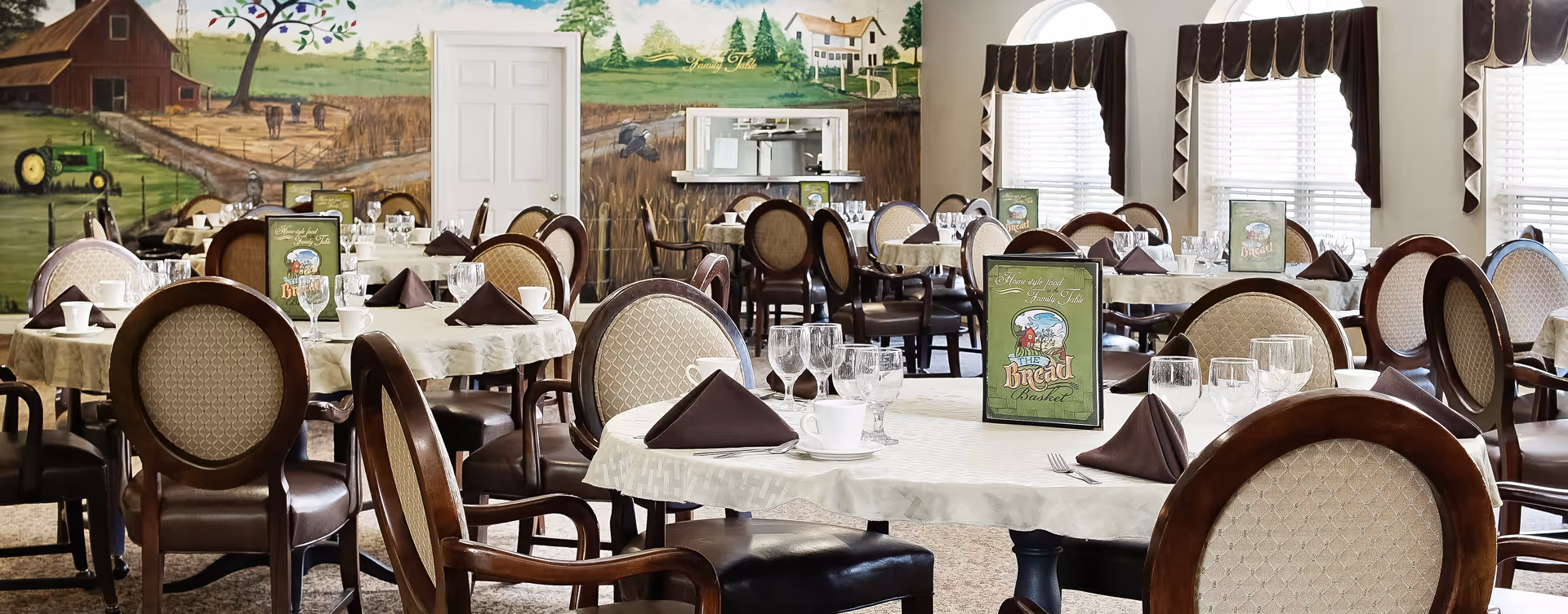 A dining room with multiple round tables covered with white tablecloths, each set with brown folded napkins, glassware, and white cups. The chairs have wooden frames with cushioned seats and backs. The walls feature a mural depicting a rural farm scene with a barn, tractor, and fields. There are windows with white blinds and dark valances on the right side of the room.