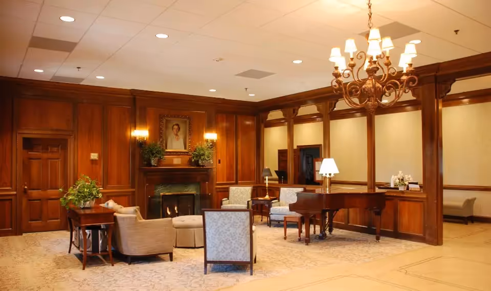 A cozy living room area with wooden panel walls, a lit fireplace, a portrait above the fireplace, several upholstered chairs and a sofa arranged around a round ottoman, a grand piano with a lamp on it, and a chandelier hanging from the ceiling.