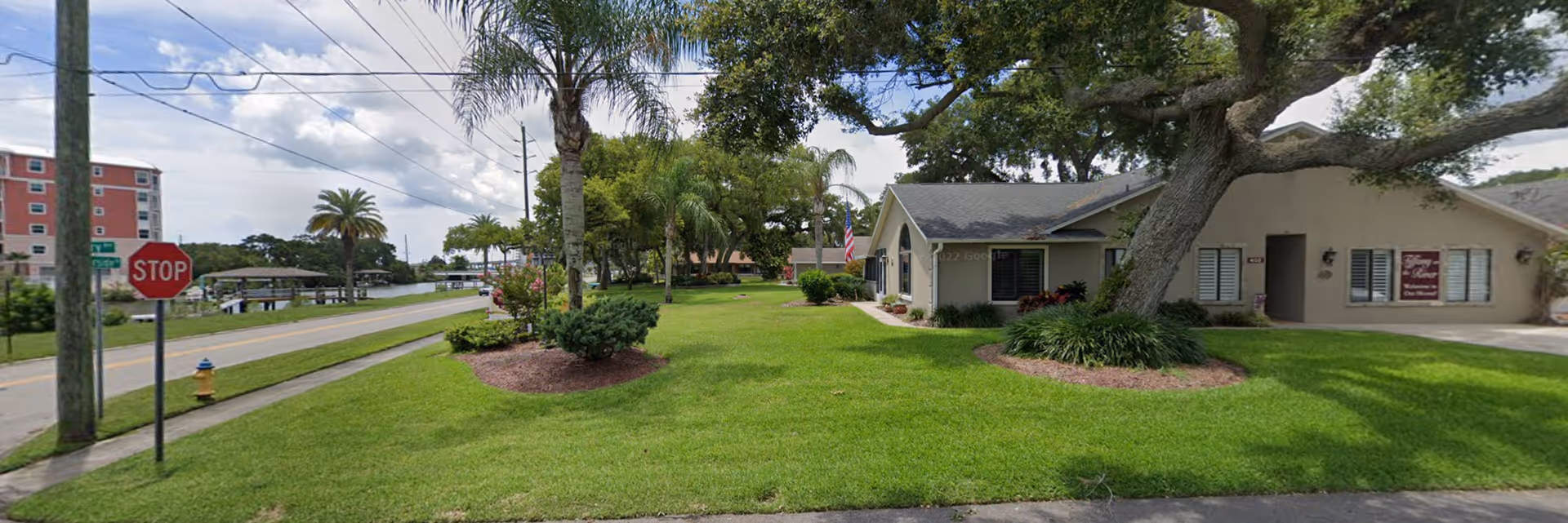 Exterior view of Riverside Care Center/Tiffany On The River showing a single-story building with a well-maintained lawn, palm trees, and a stop sign near the street. The sky is partly cloudy.