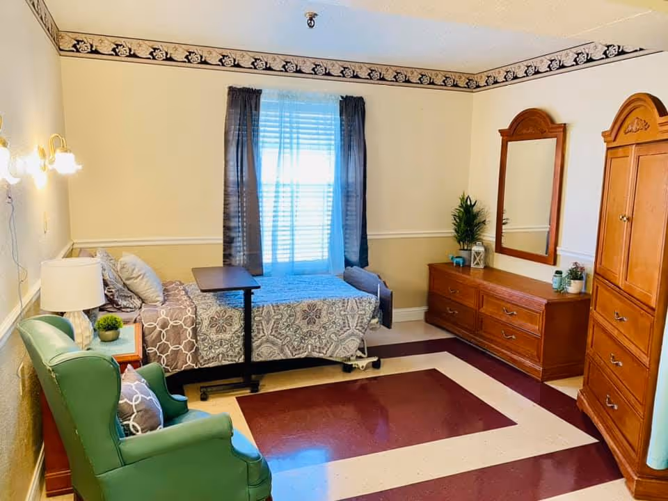 A well-lit bedroom in a senior living facility featuring a single bed with patterned bedding, a green armchair with a pillow, a wooden dresser with a mirror, a wooden armoire, a small side table with a lamp and plant, and a window with sheer and dark curtains.