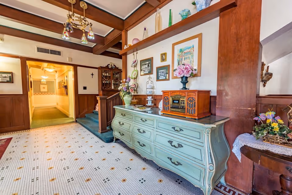 Interior view of a vintage-style hallway with a greenish-blue wooden dresser topped with a retro radio and flower arrangements. The walls are decorated with framed pictures and shelves holding decorative glass bottles. The floor has a mosaic tile pattern, and there is a wooden staircase with green carpet leading up. A chandelier with colorful stained glass shades hangs from the ceiling.