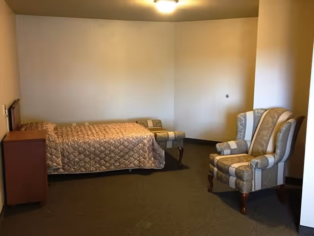 A simple bedroom with a single bed covered in a quilted bedspread, a wooden nightstand beside the bed, a striped upholstered armchair, and a matching ottoman against a plain beige wall.