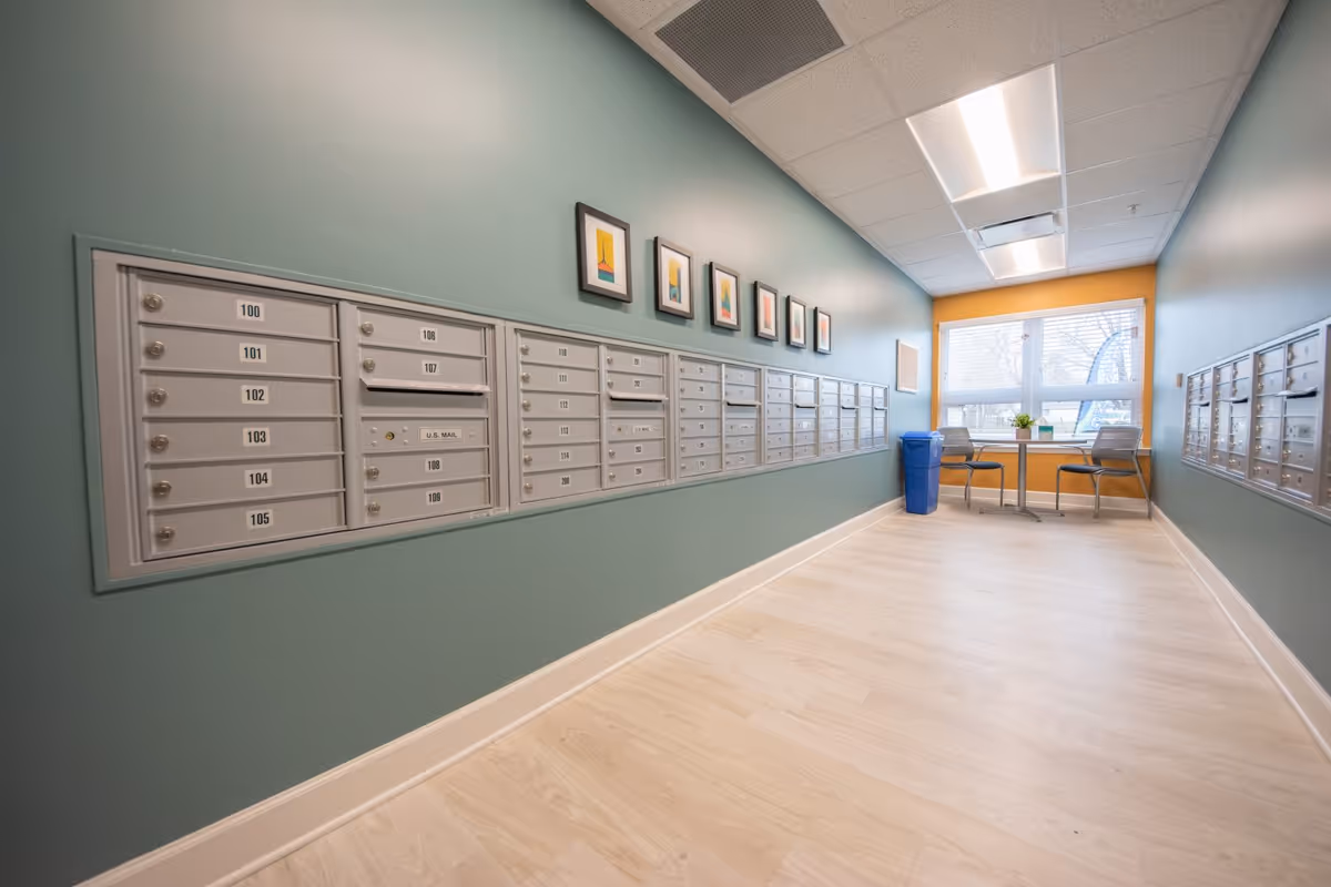 A hallway with multiple rows of mailboxes mounted on both walls. The walls are painted green with a yellow accent wall at the end. There are six framed abstract art pieces above the mailboxes on the left wall. At the end of the hallway, there is a small table with two chairs and a blue recycling bin next to it. A large window lets in natural light.