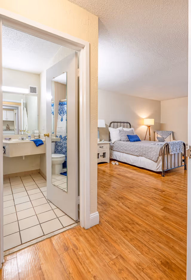 View of a bedroom with a metal bed frame, gray bedding, and blue accent pillows. A bedside table with a lamp and a chair with a throw blanket are visible. To the left, an open door reveals a bathroom with a sink, mirror, toilet, and a shower curtain with a blue and white pattern. The floor is wooden in the bedroom and tiled in the bathroom.