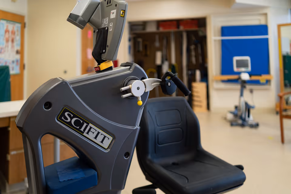 A close-up view of a SCIFIT exercise machine with a black seat in a rehabilitation or physical therapy room. In the background, there is another exercise machine, a blue padded wall mat, and various therapy equipment.