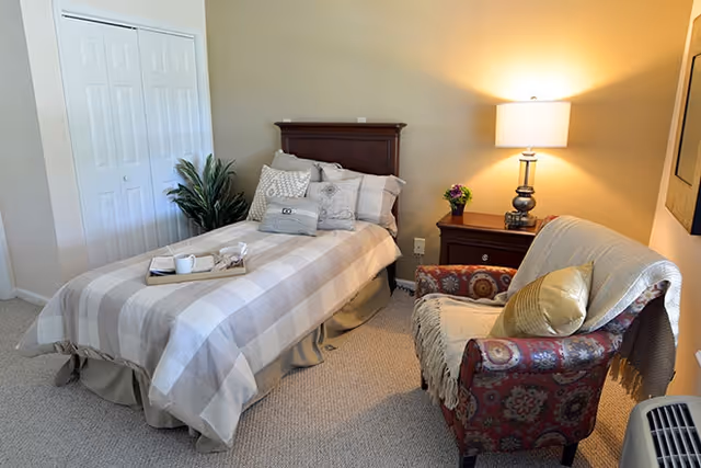 A cozy bedroom with a single bed covered in a beige and white checkered bedspread, multiple pillows, and a tray with a cup and book on the bed. Next to the bed is a wooden nightstand with a lamp and a small flower vase. An upholstered armchair with a patterned fabric and a throw blanket is positioned near the nightstand. A closet with white double doors and a green plant are visible in the background.